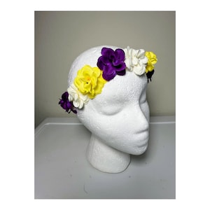 Puede incluir: Una corona floral con flores amarillas, moradas y blancas. Las flores están dispuestas en un patrón circular alrededor de una cabeza de espuma blanca. La corona es un accesorio decorativo.