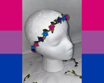 Könnte beinhalten: Ein Blumenkranz mit kleinen künstlichen Rosen in Rosa-, Blau- und Violetttönen, durchsetzt mit grünen Stoffblättern. Das Accessoire wird auf einem weißen Schaumstoff-Mannequinkopf präsentiert, wobei darunter zusätzliche grüne Ranken liegen.