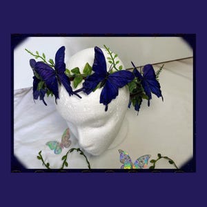 May include: A decorative headpiece featuring large, deep blue butterfly accents. The butterflies are arranged around a crown of green leaves and foliage. The headpiece is displayed on a white foam head, with additional butterfly decorations in the background.