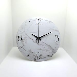 May include: A white round clock with black hands and black Roman numeral markings on a white marble background. The clock face has a white marble pattern.