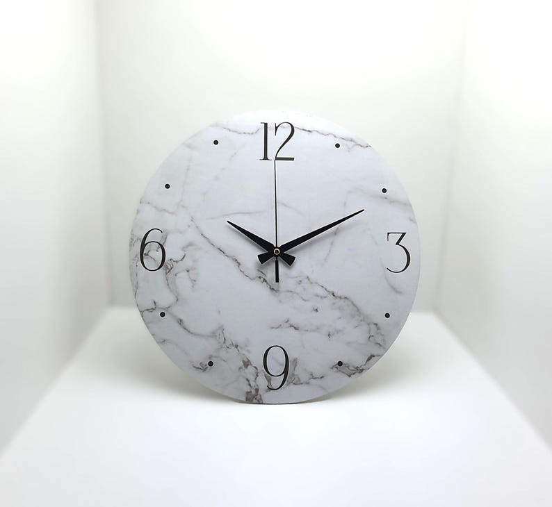 May include: A white round clock with black hands and black Roman numeral markings on a white marble background. The clock face has a white marble pattern.