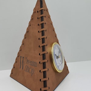 May include: A brown leather pyramid-shaped clock with a gold-colored face and black hands. The clock has a stitched design and the text "W Premium Shop" is engraved on the front.
