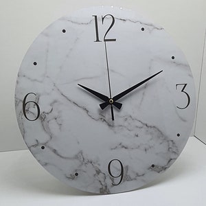 May include: A white round clock with black hands and black numbers on a white marble background. The numbers 12, 3, 6, and 9 are visible.