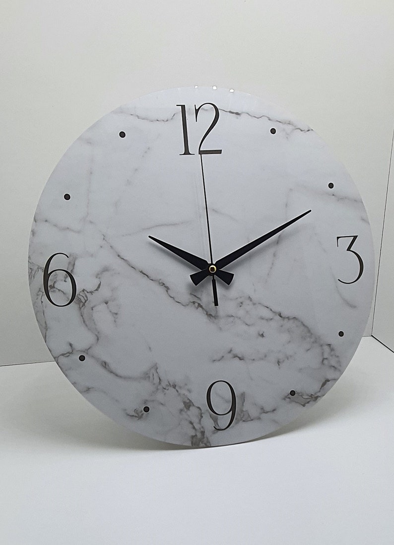 May include: A white round clock with black hands and black numbers on a white marble background. The numbers 12, 3, 6, and 9 are visible.