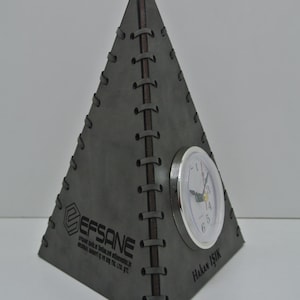 May include: A gray leather pyramid-shaped desk clock with a silver face and black hands. The clock is stitched with brown thread and has the text "EFSANE" and "Hakan IŞIK" engraved on the base.