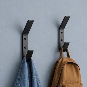 May include: Two matte black metal wall hooks mounted vertically on a light blue wall. One hook holds a blue denim jacket, while the other hook supports a tan canvas backpack. The hooks feature a dual-prong design with visible screw hardware.