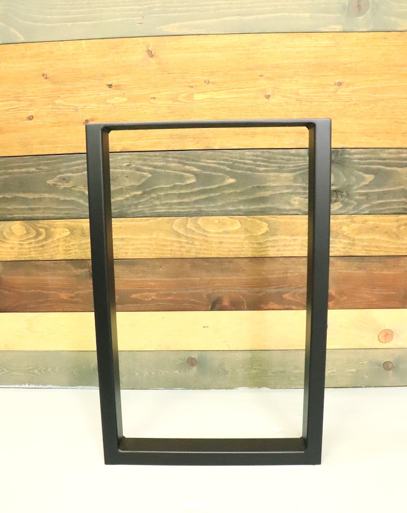 Op de afbeelding: Zwarte metalen tafelpoten met een rechthoekig frame. De poten staan op een wit oppervlak voor een achtergrond van houten panelen.