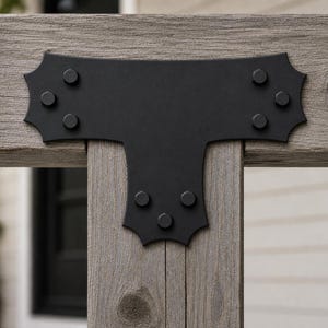May include: A matte black metal T-shaped structural connector plate featuring decorative scalloped edges and twelve circular bolt heads. The bracket is mounted onto a weathered gray wooden post and beam assembly, securing the joint connection.