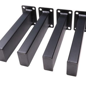 May include: Four black metal table legs. Each leg is rectangular with a square mounting plate with four holes. The legs are angled and appear to be made of steel. The legs are a matte black color.