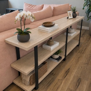 May include: A long, narrow console table with three light wood-toned shelves and a black metal frame. The table features a white orchid in a dark pot, stacked books, a wooden bowl, a small framed picture, and a woven basket on the lower shelf.