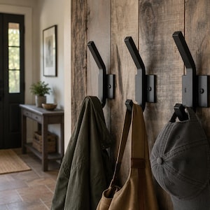 May include: Three matte black metal wall hooks mounted on a vertical wooden plank surface. Each hook features a dual-prong design with a rectangular base plate secured by two visible screws. Items including a jacket, bag, and cap hang from them.