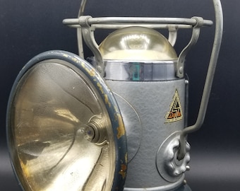 May include: Vintage gray and silver metal lantern with a large, round lens and a curved handle. The lens is open, revealing the interior. The lantern has a base and a decorative emblem.