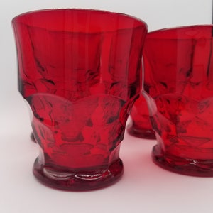 Könnte beinhalten: Vier rote Trinkgläser. Die Gläser bestehen aus dickem, strukturiertem Glas mit einem dekorativen Muster um den Boden. Die Gläser haben ein klassisches, Vintage-Aussehen.