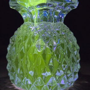 Op de afbeelding: Een heldere glazen vaas in de vorm van een ananas met een getextureerd oppervlak. De vaas gloeit groen onder een blacklight.