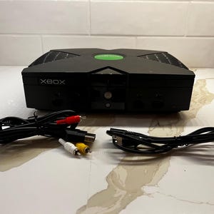 May include: Black Xbox video game console with a green power button and the word "XBOX" printed on the front. Includes black cables with red, white, and yellow connectors.