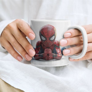 K&ouml;nnte beinhalten: Wei&szlig;e Keramiktasse mit einem detaillierten Bild von Spider-Man in seinem roten und blauen Anzug. Der Superheld wird in einer meditativen Pose dargestellt. Die Tasse hat einen gro&szlig;en, gebogenen Henkel. Die Tasse wird von einer Person gehalten.