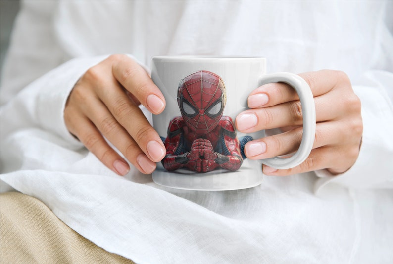 K&ouml;nnte beinhalten: Wei&szlig;e Keramiktasse mit einem detaillierten Bild von Spider-Man in seinem roten und blauen Anzug. Der Superheld wird in einer meditativen Pose dargestellt. Die Tasse hat einen gro&szlig;en, gebogenen Henkel. Die Tasse wird von einer Person gehalten.