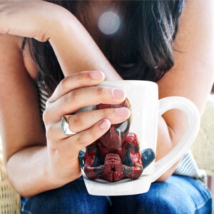 K&ouml;nnte beinhalten: Wei&szlig;e Keramik-Tasse mit detailliertem Bild von Spider-Man in meditativer Pose. Der Superheld ist in seinem klassischen rot-schwarzen Anzug dargestellt, die H&auml;nde gefaltet. Die Tasse hat einen gebogenen Henkel.