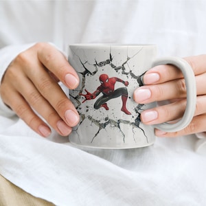Puede incluir: Una taza de cer&aacute;mica blanca con un dise&ntilde;o de pared agrietada que presenta un personaje de Spiderman rojo y azul rompiendo la pared.