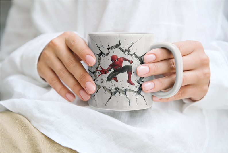 Puede incluir: Una taza de cer&aacute;mica blanca con un dise&ntilde;o de pared agrietada que presenta un personaje de Spiderman rojo y azul rompiendo la pared.