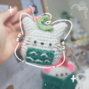 May include: White crocheted cat keychain with green leaf detail and a green and white striped scarf. The cat has black eyes and whiskers.