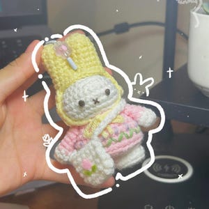 May include: A handmade crochet bunny keychain in white, yellow, and pink. The bunny wears a yellow hat, pink sweater, and a white bag with a flower. The keychain has a small charm and is outlined with a white border.