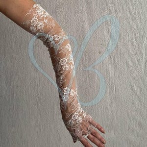 K&ouml;nnte beinhalten: Wei&szlig;e Spitzenhandschuhe mit floralen Mustern. Die Handschuhe reichen von den Fingerspitzen bis zum Oberarm. Das zarte Spitzendesign ist auf der Haut sichtbar und erzeugt einen eleganten und romantischen Look.
