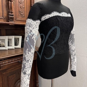 May include: A black long-sleeved top with a lace overlay. The lace is white and has a floral pattern. The top has a high neck and a fitted bodice.