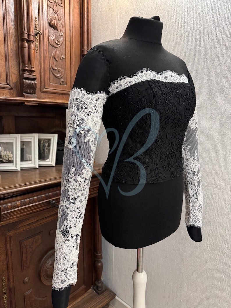 May include: A black long-sleeved top with a lace overlay. The lace is white and has a floral pattern. The top has a high neck and a fitted bodice.