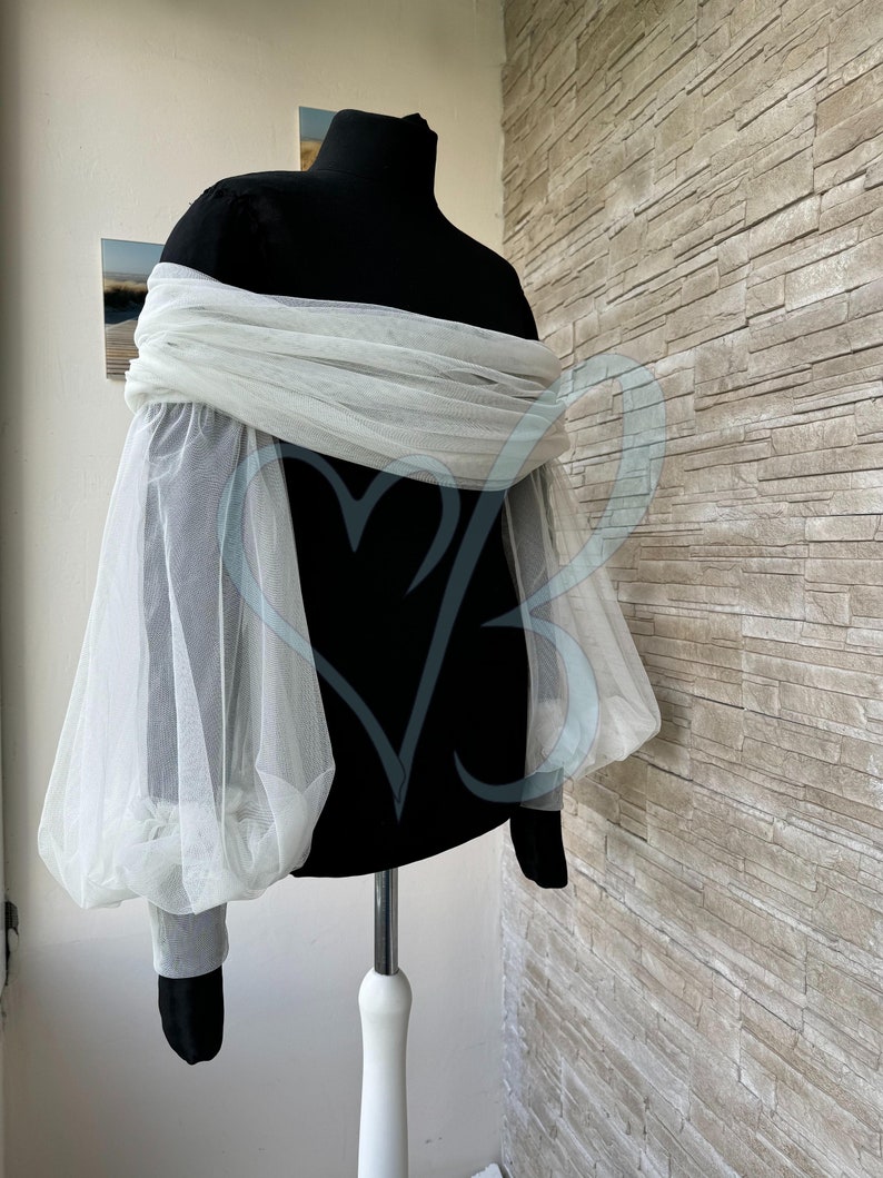 May include: A white tulle off-the-shoulder top with long, puffy sleeves. The top is on a black mannequin.