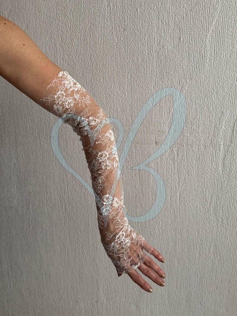 K&ouml;nnte beinhalten: Wei&szlig;e Spitzenhandschuhe mit floralen Mustern. Die Handschuhe reichen von den Fingerspitzen bis zum Oberarm. Das zarte Spitzendesign ist auf der Haut sichtbar und erzeugt einen eleganten und romantischen Look.