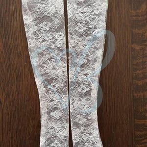 K&ouml;nnte beinhalten: Zwei wei&szlig;e Armstulpen aus Spitze mit floralem Muster werden auf einer dunklen Holzoberfl&auml;che gezeigt. Der zarte Spitzenstoff ist aufwendig detailliert und schafft eine romantische und elegante &Auml;sthetik. Diese Armstulpen sind ein stilvolles Accessoire.