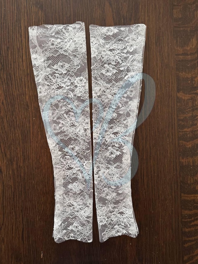 K&ouml;nnte beinhalten: Zwei wei&szlig;e Armstulpen aus Spitze mit floralem Muster werden auf einer dunklen Holzoberfl&auml;che gezeigt. Der zarte Spitzenstoff ist aufwendig detailliert und schafft eine romantische und elegante &Auml;sthetik. Diese Armstulpen sind ein stilvolles Accessoire.