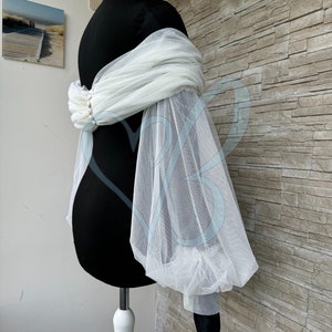 May include: White tulle bridal shrug with puffed sleeves and pearl embellishments. The shrug is draped over a black dress form.