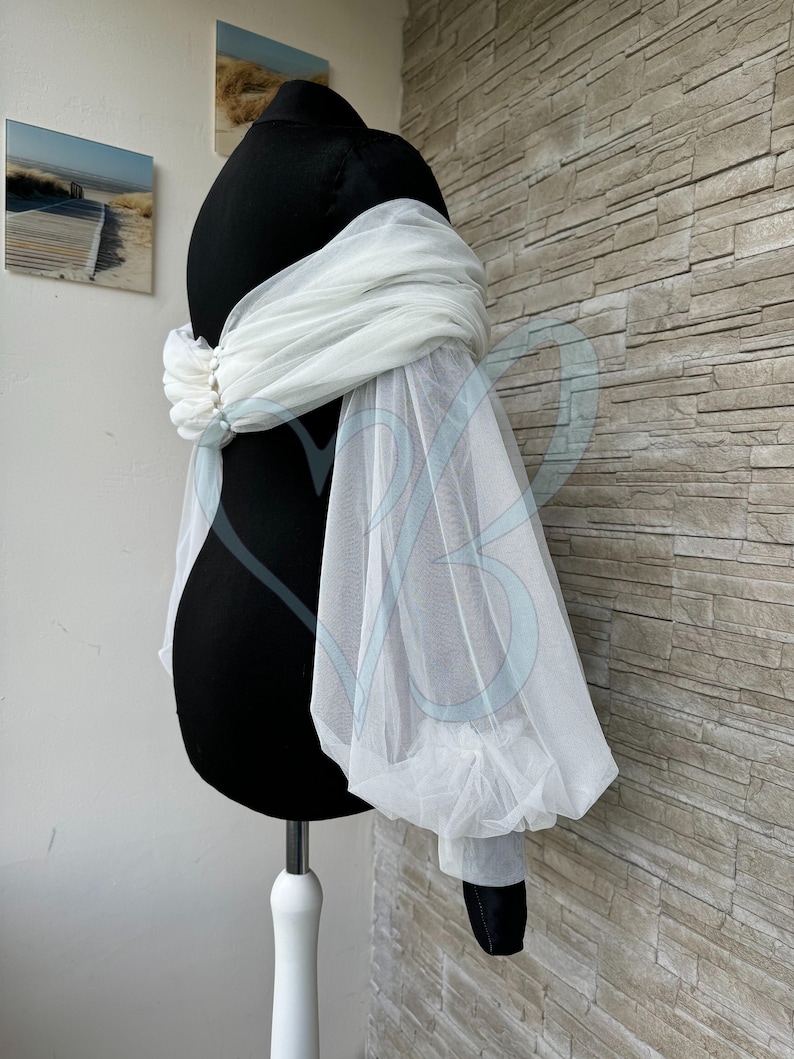 May include: White tulle bridal shrug with puffed sleeves and pearl embellishments. The shrug is draped over a black dress form.
