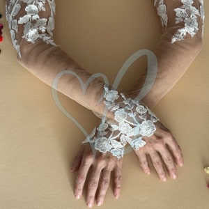 May include: A pair of sheer white lace gloves with floral embroidery. The gloves are worn on a person's arms, with the hands crossed in front of them.