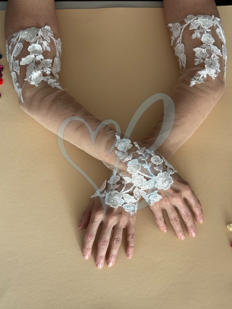 May include: A pair of sheer white lace gloves with floral embroidery. The gloves are worn on a person's arms, with the hands crossed in front of them.
