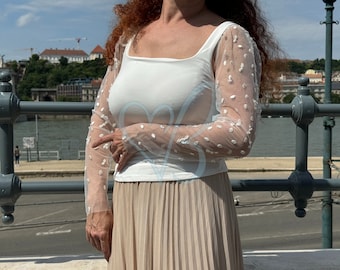 May include: A white top with a scoop neck and long sleeves. The sleeves are made of a sheer fabric with white polka dots. The top is paired with a beige pleated skirt.