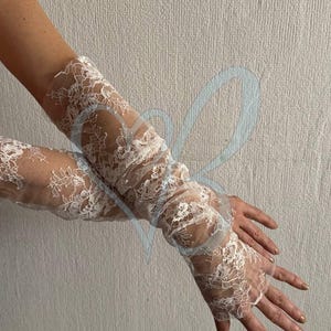 K&ouml;nnte beinhalten: Wei&szlig;e Spitzengloves mit floralem Muster. Die fingerlosen Handschuhe reichen den Arm hinauf und zeigen filigrane Spitze. Der Look ist romantisch und elegant. Der Hintergrund ist eine schlichte, hellfarbige Wand.