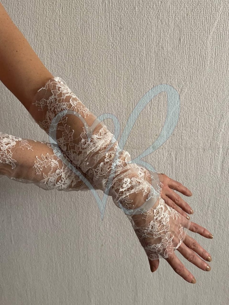 K&ouml;nnte beinhalten: Wei&szlig;e Spitzengloves mit floralem Muster. Die fingerlosen Handschuhe reichen den Arm hinauf und zeigen filigrane Spitze. Der Look ist romantisch und elegant. Der Hintergrund ist eine schlichte, hellfarbige Wand.