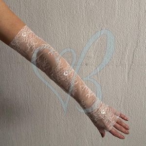 May include: Ivory lace fingerless gloves. The gloves are long, extending up the arm, and feature a delicate floral lace pattern. Small sequins are scattered across the lace, adding a touch of sparkle. The background is a neutral, textured wall.
