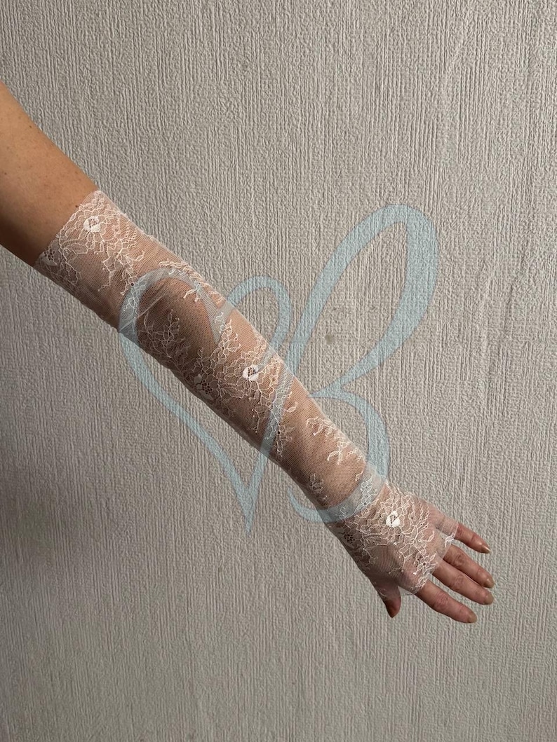 May include: Ivory lace fingerless gloves. The gloves are long, extending up the arm, and feature a delicate floral lace pattern. Small sequins are scattered across the lace, adding a touch of sparkle. The background is a neutral, textured wall.