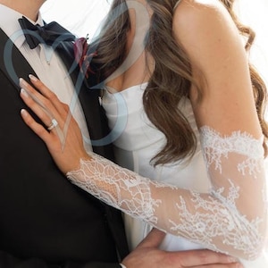 May include: A bride's white lace sleeves and a wedding ring are visible. The sleeves are long and fitted, with intricate floral lace patterns. The bride is embracing a person wearing a black tuxedo with a bow tie.