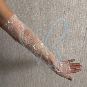 May include: Elegant white lace gloves, extending from the fingertips to the upper arm. The gloves feature a delicate floral pattern and are made of a sheer, lightweight fabric. Perfect for weddings or formal events.