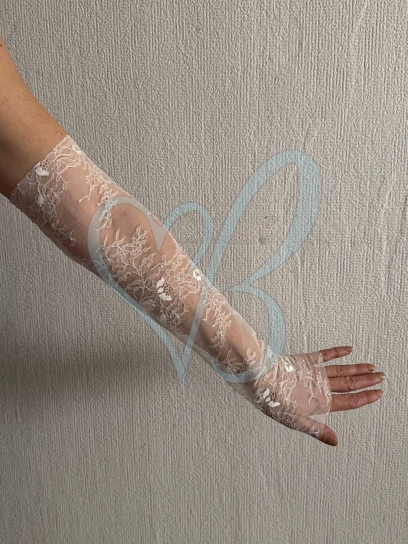 May include: Elegant white lace gloves, extending from the fingertips to the upper arm. The gloves feature a delicate floral pattern and are made of a sheer, lightweight fabric. Perfect for weddings or formal events.