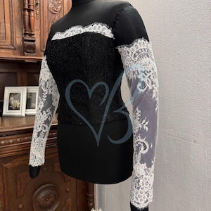 May include: A black top with long sleeves featuring white lace detailing. The lace is intricate and delicate, with a floral pattern. The top has a high neckline and a fitted silhouette.