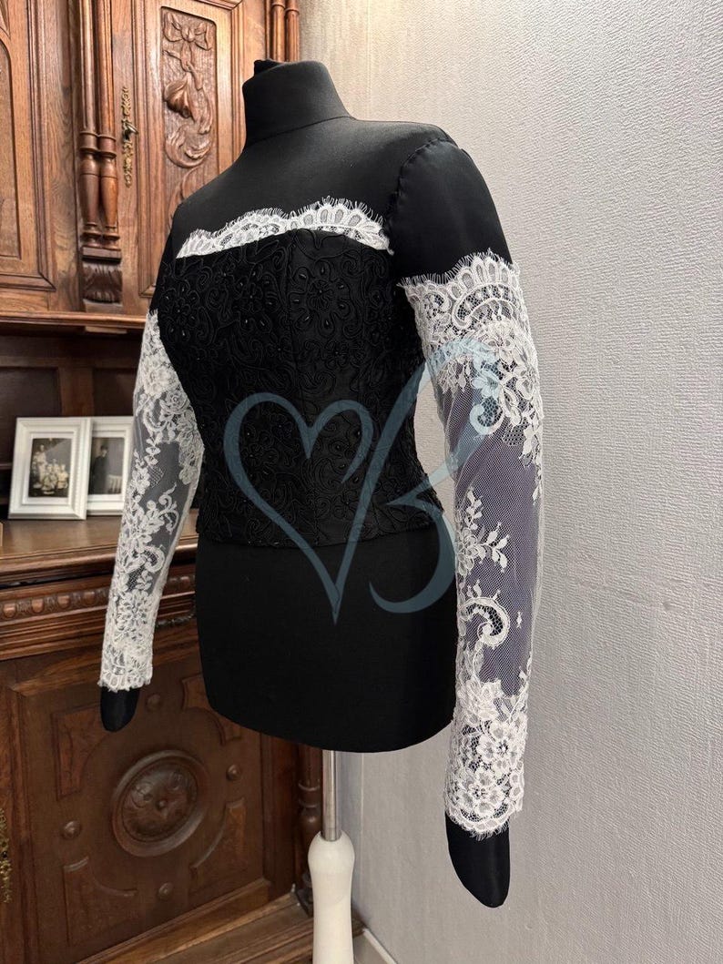 May include: A black top with long sleeves featuring white lace detailing. The lace is intricate and delicate, with a floral pattern. The top has a high neckline and a fitted silhouette.