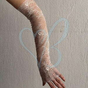 May include: Ivory lace gloves with a delicate floral pattern. The gloves extend from the fingertips to the upper arm. The sheer fabric is adorned with small, sparkling embellishments, adding a touch of elegance. Perfect for special occasions.