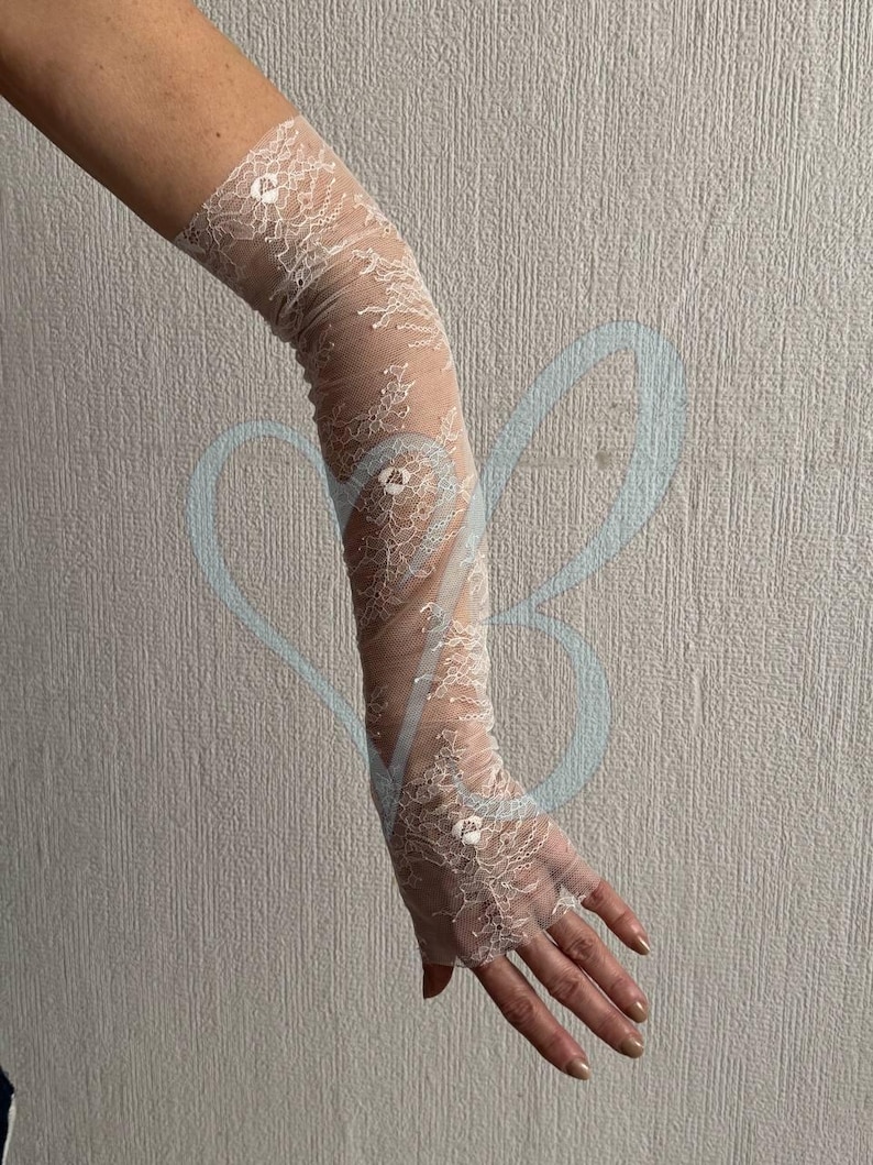 May include: Ivory lace gloves with a delicate floral pattern. The gloves extend from the fingertips to the upper arm. The sheer fabric is adorned with small, sparkling embellishments, adding a touch of elegance. Perfect for special occasions.