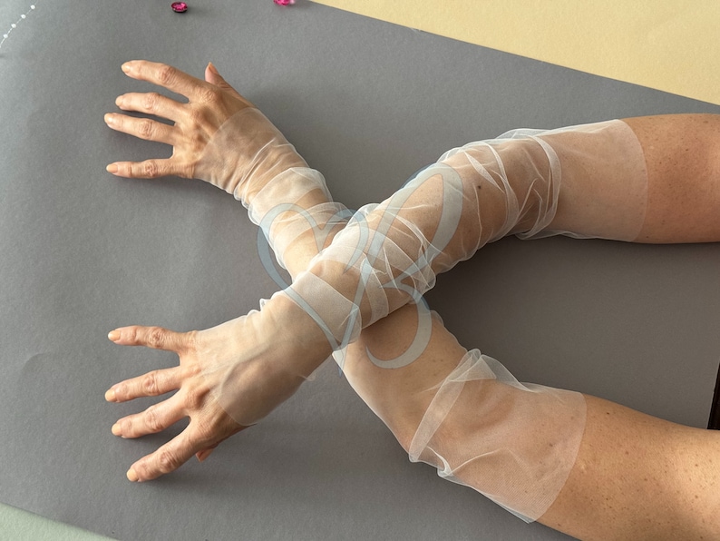 K&ouml;nnte beinhalten: Ein Paar wei&szlig;e, durchsichtige Ellbogenhandschuhe. Die Handschuhe sind aus einem zarten, leichten Stoff gefertigt und perfekt, um jedem Outfit einen Hauch von Eleganz zu verleihen.